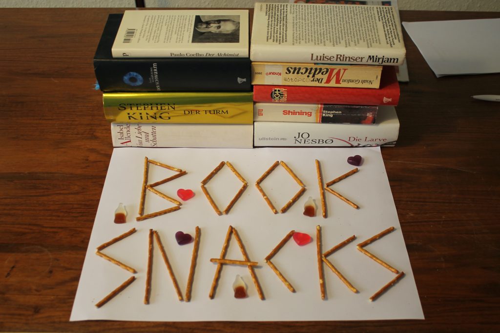 Vor einem Stapel Büchern ist auf dem Tisch aus Salzstangen zusammengesetzt das Wort BOOKSNACKS zu lesen.